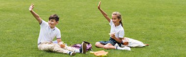 Positive multiethnic schoolkids holding sandwiches near backpacks on grass in park, banner  clipart