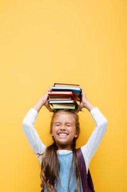 Happy schoolgirl holding books on head isolated on yellow  clipart