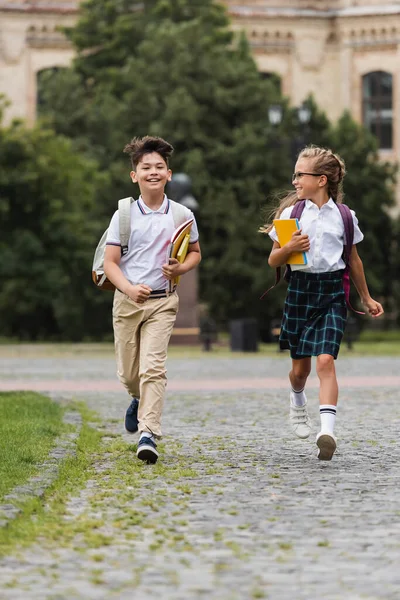 Positive multiethnic pupils with backpacks running on walkway outdoors