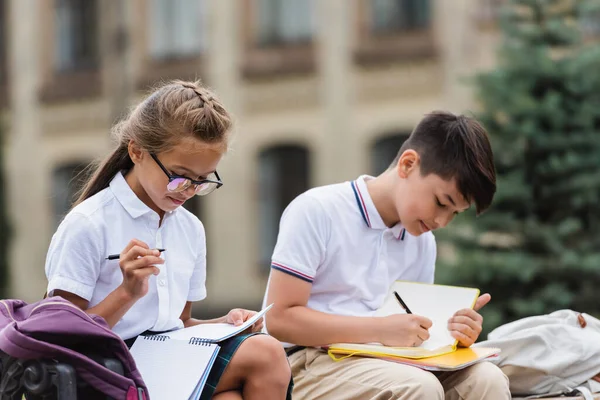 Preteen Interraciale Schoolkinderen Schrijven Notitieboekjes Buurt Van Rugzakken Buiten — Stockfoto