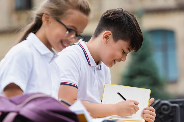 Smiling Asian Schoolboy Writing Notebook Blurred Friend Outdoors — Stok Foto