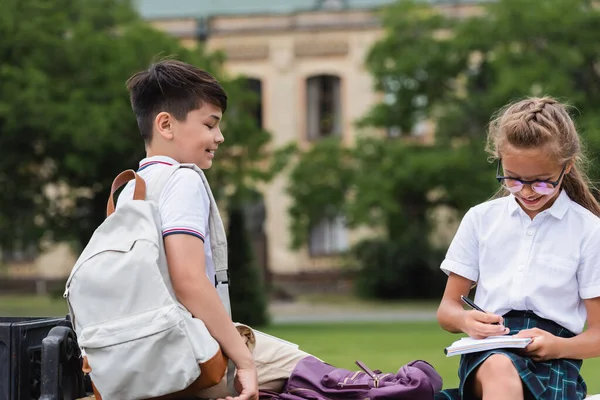 Positivo Estudante Escrevendo Notebook Perto Asiático Amigo Com Mochila Banco — Fotografia de Stock