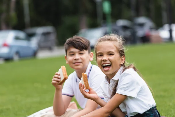 Spännande Skolflicka Håller Smörgås Nära Suddig Asiatisk Klasskamrat Parken — Stockfoto