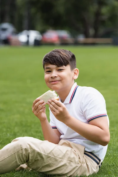 Gülümseyen Asyalı Okul Çocuğu Parkta Çimlerin Üzerinde Sandviç Tutuyor — Stok fotoğraf