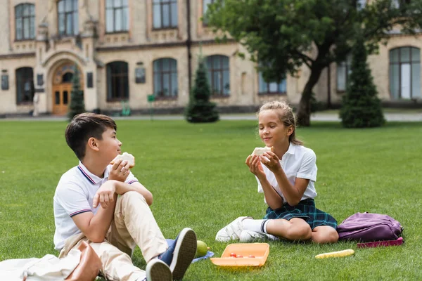 Colegiales Multiétnicos Sosteniendo Sándwiches Cerca Mochilas Hierba Parque — Foto de Stock