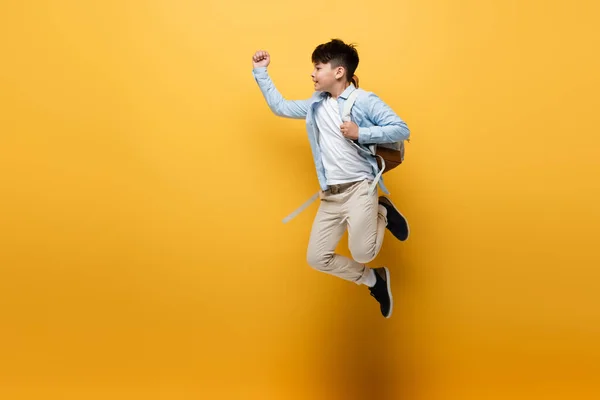 Side view of asian schoolkid with backpack jumping on yellow background