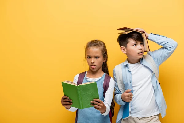 Colegiala Leyendo Libro Sobresaliendo Lengua Cerca Asiático Amigo Aislado Amarillo —  Fotos de Stock