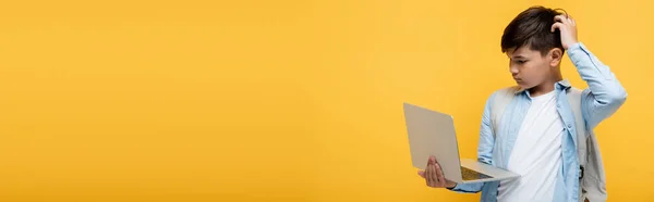 Pensive asian pupil with backpack looking at laptop isolated on yellow, banner