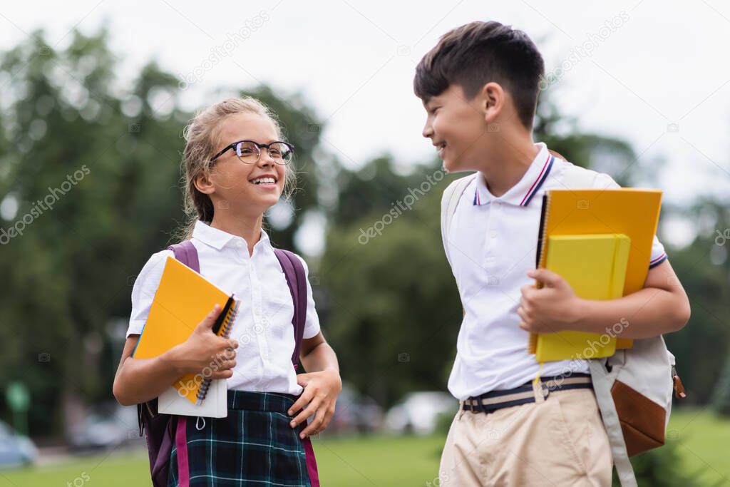 Smiling pupil in eyeglasses holding notebooks near asian friend in park 