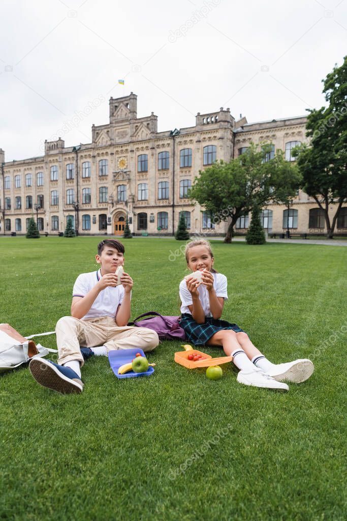 Positive preteen schoolkids holding sandwiches near lunchboxes on grass in park 