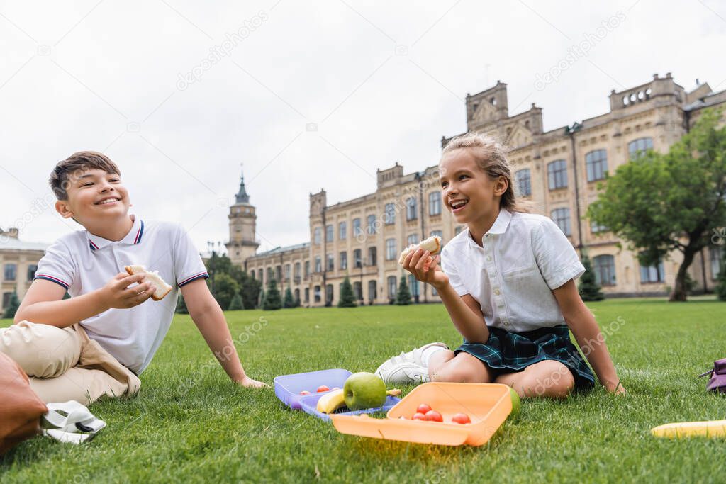 Positive multiethnic kids holding sandwiches near lunchboxes on lawn 