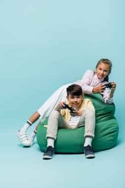 KYIV, UKRAINE - 2 Temmuz 2021: Mavi arka planda puf koltukta video oyunu oynayan çok ırklı çocuklar 