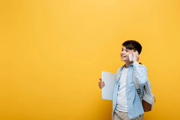 Sonriente Asiático Colegial Hablando Smartphone Sosteniendo Portátil Aislado Amarillo —  Fotos de Stock