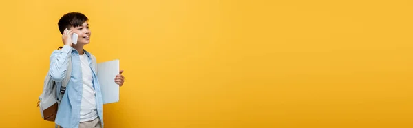 Позитивний Азіатський Школяр Розмовляє Смартфоні Тримає Ноутбук Ізольований Жовтому Банер — стокове фото