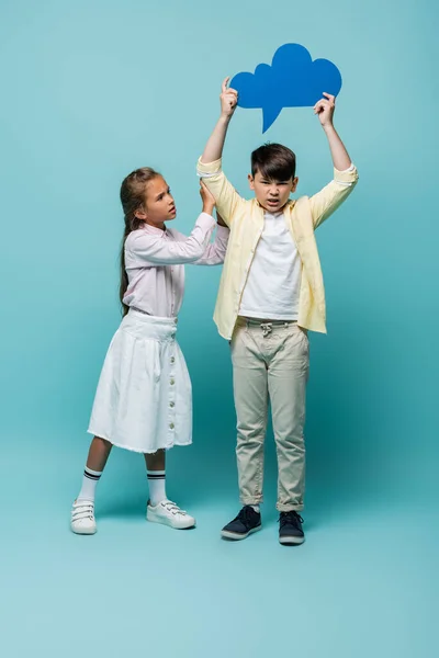 Schoolgirl looking at angry asian friend with thought bubble on blue background