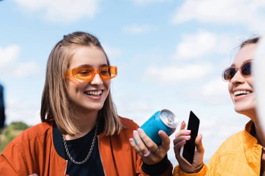 Soda kutusu olan genç kadınlar ve açık havada gülümseyen boş ekranlı akıllı telefon.