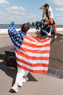 Neşeli çoklu etnik arkadaşlar paten parkında Amerikan bayrağı taşıyan kadını işaret ediyorlar.