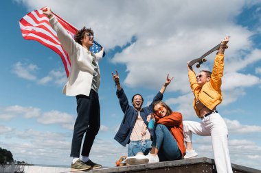 Heyecanlı Asyalı adam Amerika bayrağı ve kaykayıyla arkadaşlarının yanında kaya işaretleri gösteriyor.
