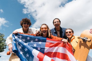 Heyecanlı ırklar arası patencilerin düşük açılı görüntüsü ABD bayrağıyla mavi ve bulutlu gökyüzünün altında kameraya bakıyor