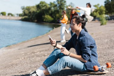 asian man in eyeglasses using mobile phone while sitting on longboard near river and blurred friends clipart