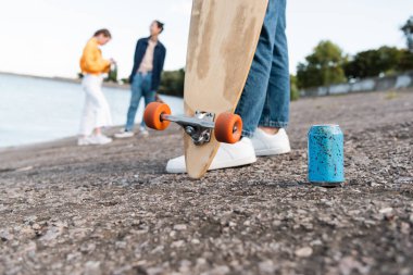 partial view of skater with longboard near soda can and blurred friends on river bank clipart