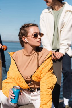 Soda kutusu olan trend bir kadın bulanık arka planda arkadaşının yanından uzağa bakıyor.