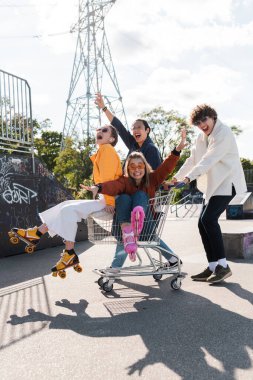 astonished multicultural friends shouting while having fun with shopping trolley in skate park clipart
