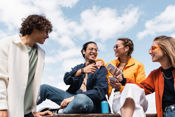 cheerful asian man pointing with finger at friends with smartphones outdoors