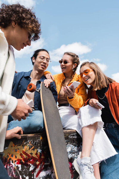 young asian man with longboard sitting with cheerful and stylish friends outdoors