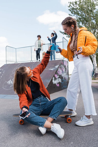 stylish women clinking soda cans near friends on skate ramp on blurred background