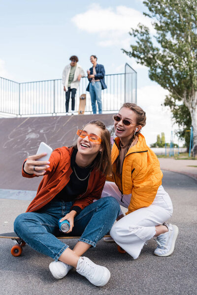 joyful women taking selfie in skate park near friends on ramp on blurred background