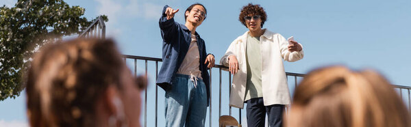 smiling asian man pointing with finger on blurred friends, banner