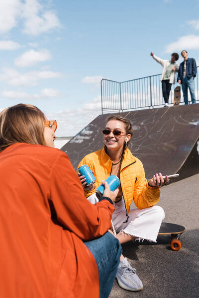 smiling woman in eyeglasses talking to friend near blurred skaters on ramp in skate park