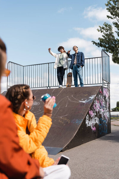 interracial skateboarders on skate ramp waving hands to young women on blurred foreground