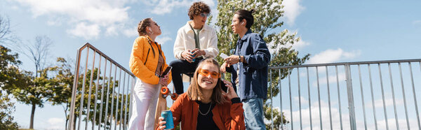 happy woman with soda can looking at camera near multicultural friends outdoors, banner