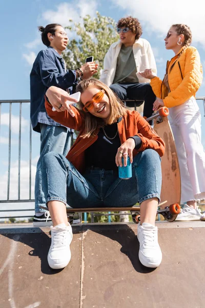 trendy woman with soda can showing victory sign while sitting on skate ramp near blurred interracial friends
