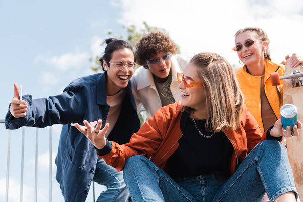 excited asian man showing thumb up near cheerful friend with soda can