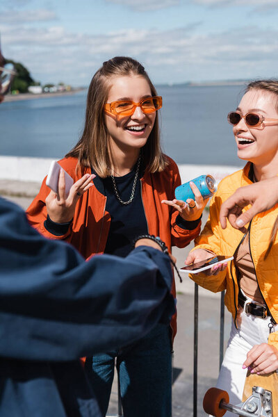 cheerful woman with smartphone and soda can talking to friends outdoors