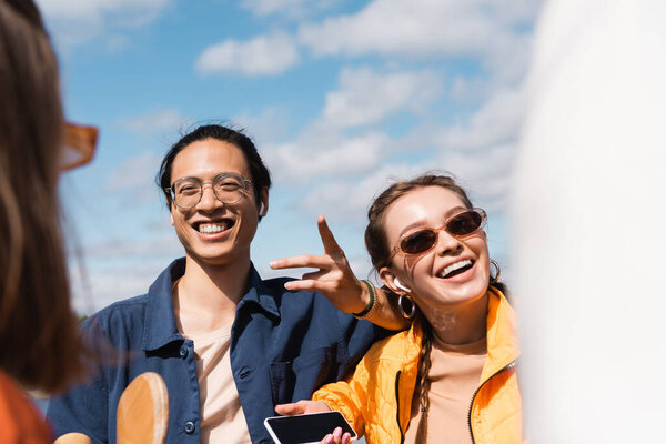 cheerful asian man pointing with finger near smiling woman in stylish sunglasses 