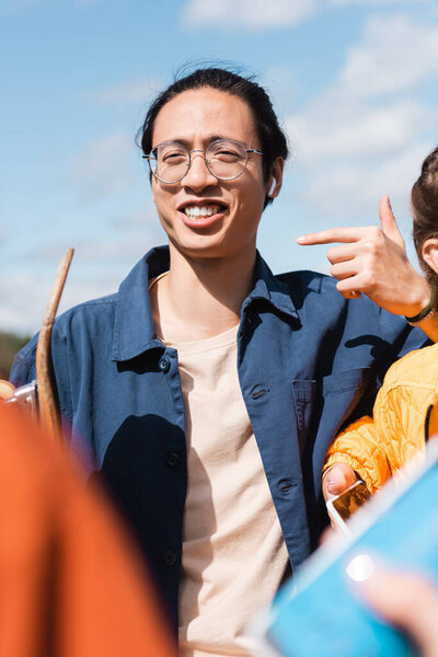 happy asian man in eyeglasses pointing with finger near blurred friends outdoors