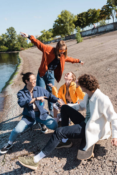 cheerful woman with outstretched hands standing near multiethnic friends on embankment