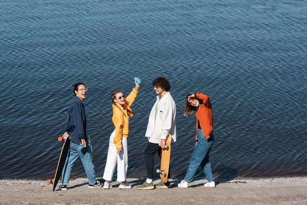 full length of happy woman with soda can in raised hand near multiethnic friends on river bank