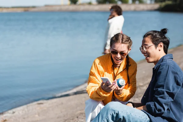 cheerful woman with soda can showing smartphone to asian friend near river