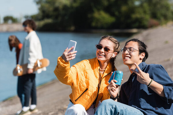 happy interracial friends clinking soda cans and taking selfie on river bank 