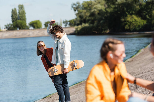 cheerful woman in sunglasses smiling near friend with skateboard on riverside