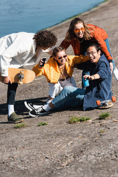 cheerful multicultural skaters with soda can and smartphone having fun on embankment