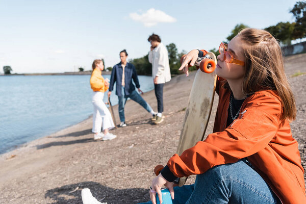 young woman in eyeglasses sitting on river bank with penny board near friends on blurred background