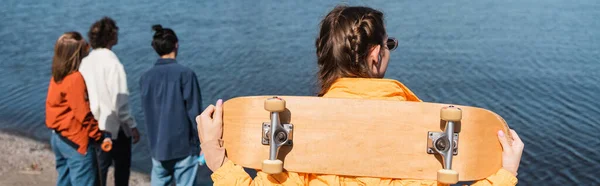 友人や川の近くにスケートボードを持つ女性のバックビュー バナー — ストック写真