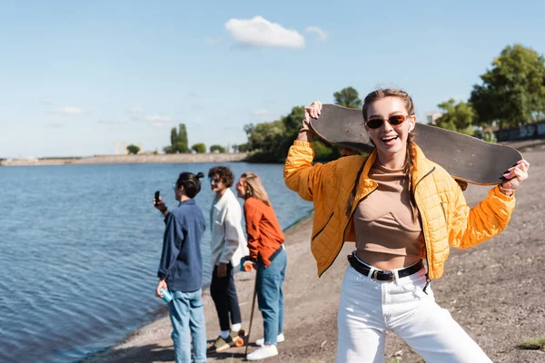 Excited Woman Skateboard Looking Camera Blurred Friends River Bank — Stock Photo, Image