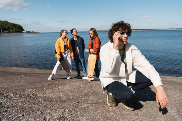 young man adjusting stylish sunglasses near interracial skaters on river bank